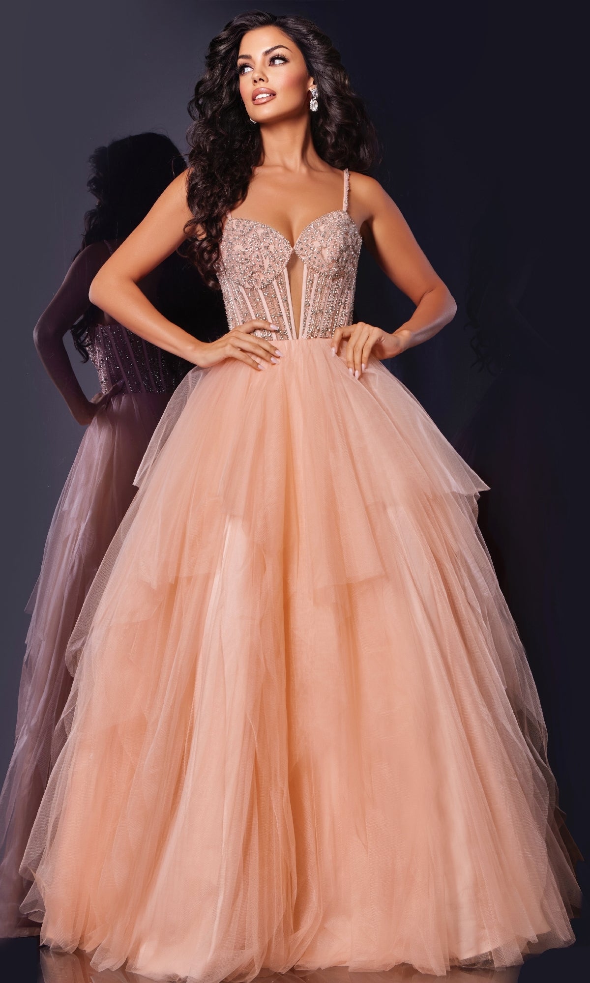A woman poses against a dark background wearing the Jovani 43254 Long Prom Dress, featuring a blush pink hue, an embellished corset bodice, and a layered tulle skirt.
